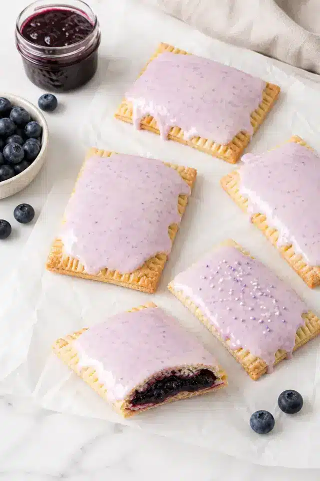 Homemade blueberry Pop Tarts on parchment with pale purple icing, blueberries, and one pastry showing filling
