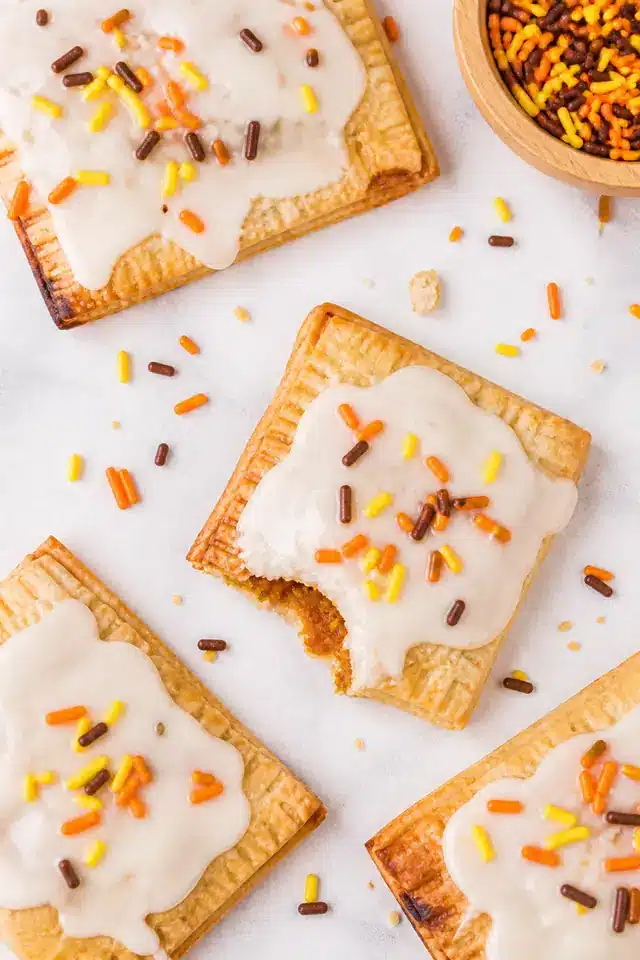 Homemade pumpkin pie pop tarts arranged on parchment with maple glaze and orange yellow and brown sprinkles