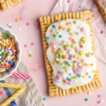 Homemade Birthday Pop Tarts 11 Homemade birthday cake Pop-Tarts topped with white icing and colorful confetti sprinkles on a pink background, surrounded by a bowl of sprinkles and birthday candles